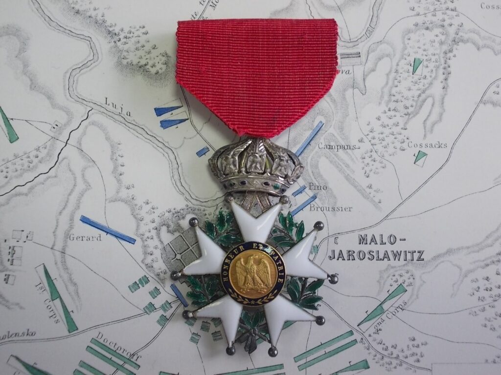 Croce di cavaliere della Legion d'Onore, modello imperiale: stella a cinque punte biforcate smaltata di bianco entro corona di alloro e quercia, sormontata dalla corona imperiale; al centro il profilo di Napoleone con la legenda «NAPOLEON EMP. DES FRANÇAIS»; nastro rosso.